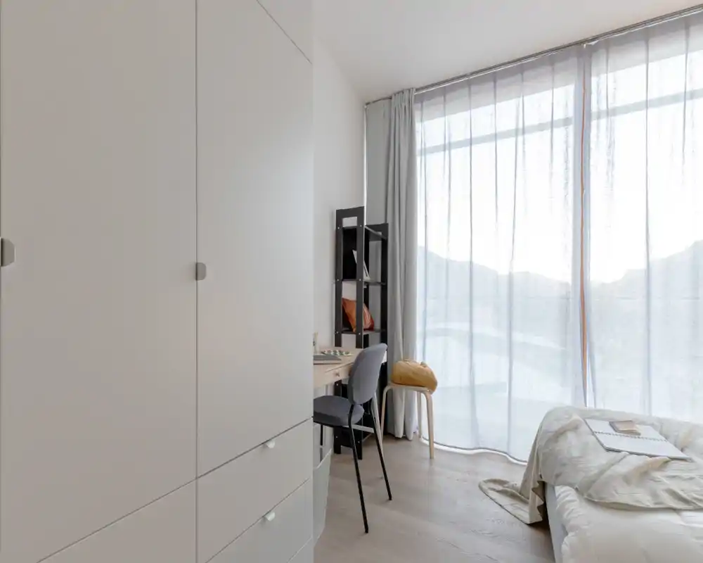 Dormitorio moderno y luminoso con cama arreglada, un pequeño escritorio y una gran ventana con cortinas translúcidas que inundan la estancia de luz natural.
