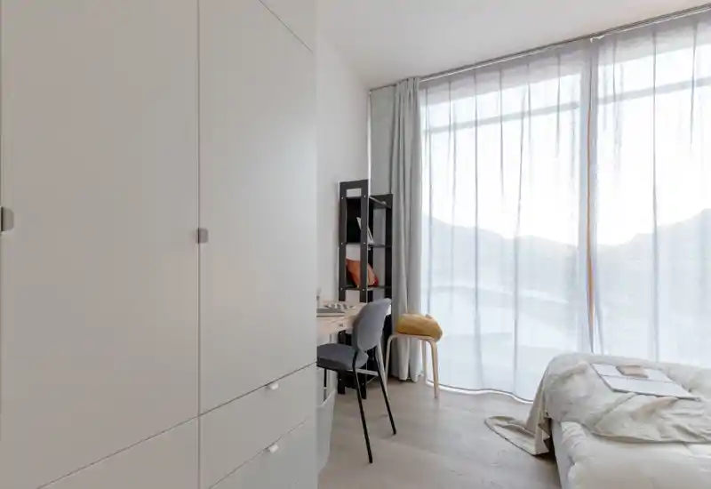 Dormitorio moderno y luminoso con cama arreglada, un pequeño escritorio y una gran ventana con cortinas translúcidas que inundan la estancia de luz natural.