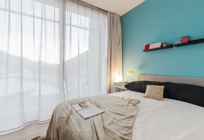 Dormitorio moderno y luminoso con cama grande en primer plano, ropa de cama suave, mesita con lámpara y estantes decorativos sobre una pared turquesa.