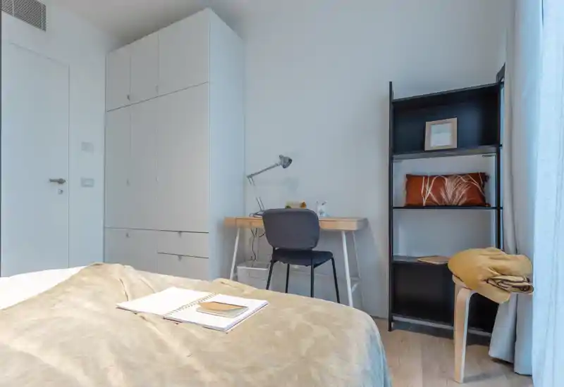 Dormitorio moderno y luminoso con la cama en primer plano, un escritorio pequeño con lámpara y silla, un armario alto y una estantería junto a la ventana.