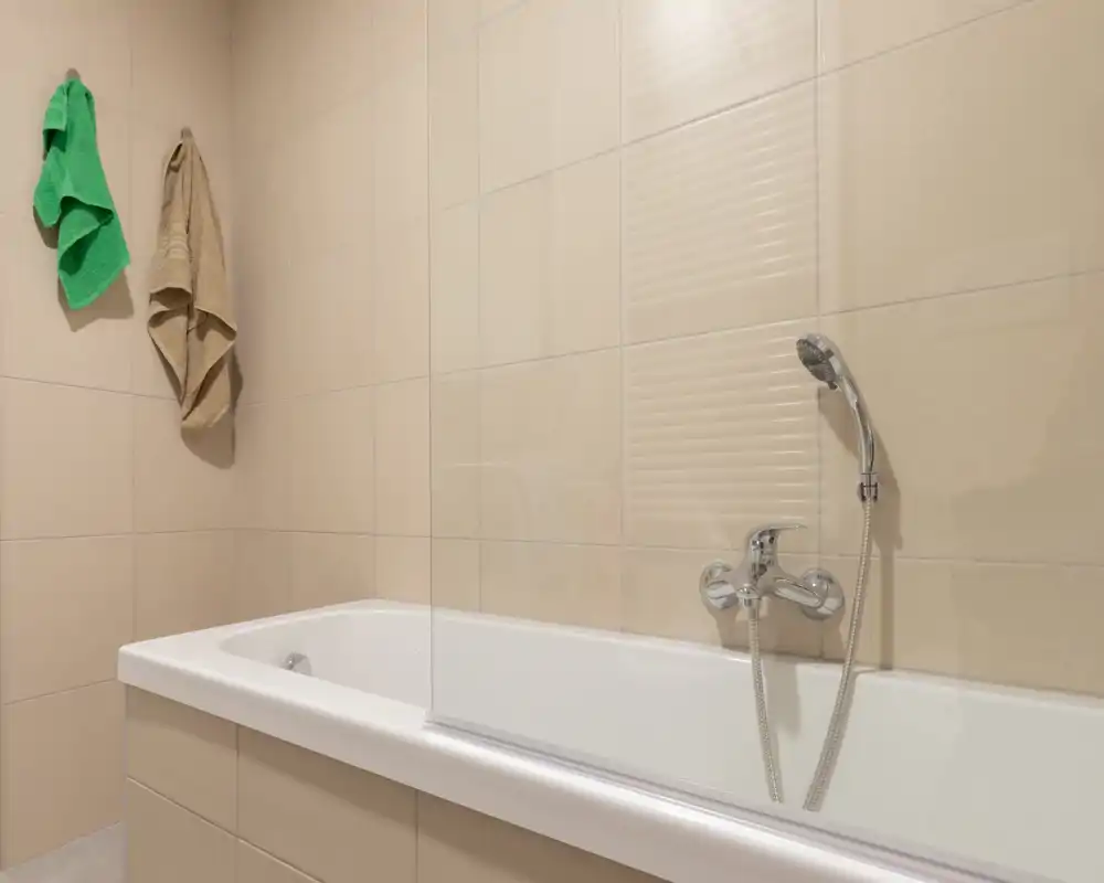 Baño de tonos neutros con bañera, ducha de mano y mampara de vidrio, paredes con azulejos beige y dos toallas colgadas.