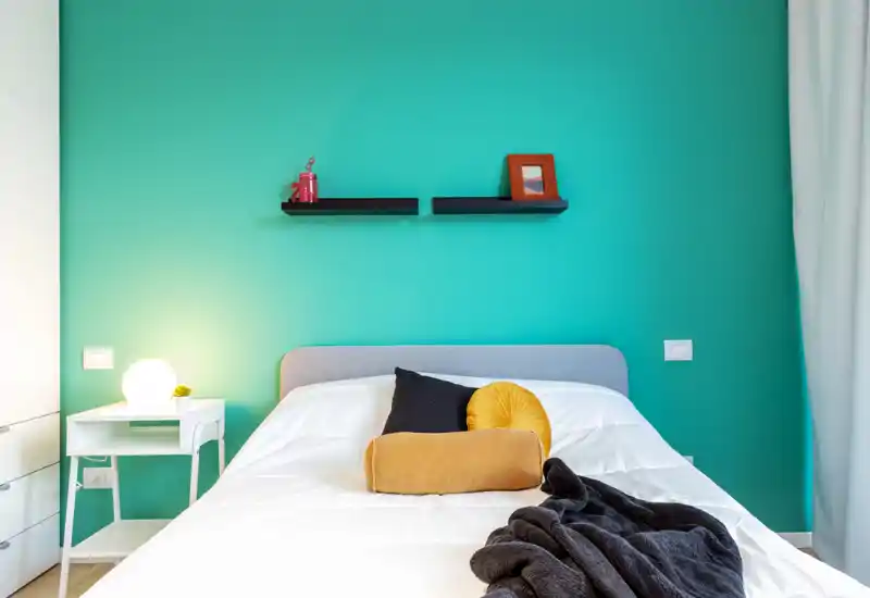 Bright, modern bedroom with a teal accent wall, neatly made double bed, decorative cushions and a soft throw. Simple nightstand with lamp and minimal floating shelves create a clean, welcoming atmosphere.