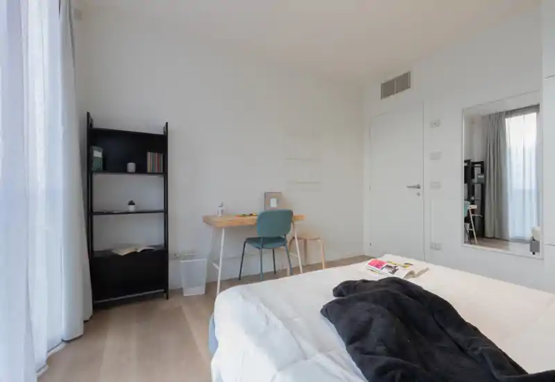 Bright, minimalist bedroom with a comfortable bed in the foreground, a small desk and chair by the wall, and a tall shelving unit near the window.