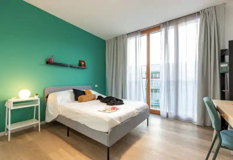 Bright, modern bedroom with a comfortable double bed against a teal accent wall, bedside lamp and large floor-to-ceiling window letting in abundant natural light.