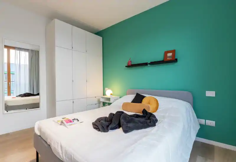Bright, styled bedroom with a comfortable double bed against a teal accent wall, wardrobe and bedside lamp; modern, welcoming space ideal for rentals.