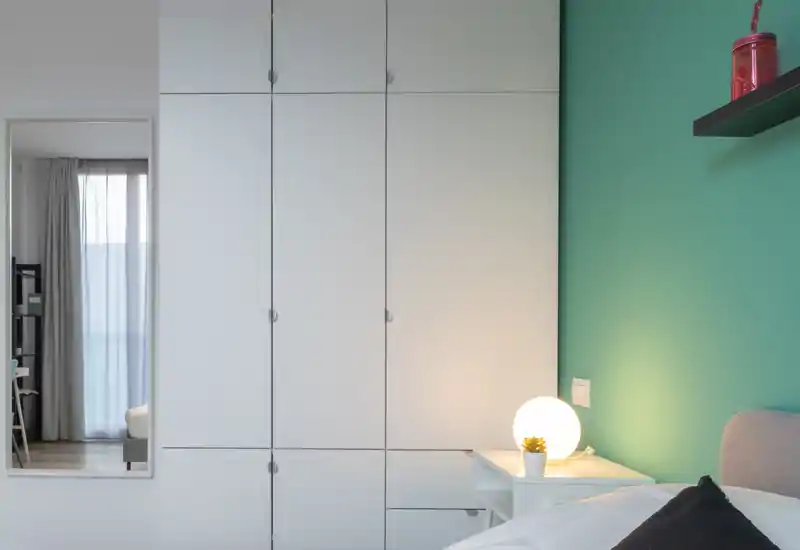 Bright, modern bedroom corner with a white wardrobe, bedside table and a cozy lamp against a teal accent wall.