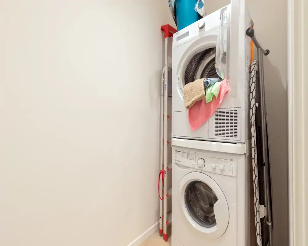 Pequeño cuarto de lavandería con lavadora y secadora apiladas y tabla de planchar; espacio práctico ideal para apartamentos eficientes.
