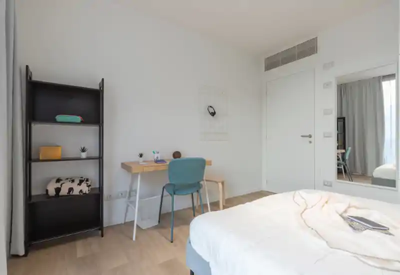 Dormitorio moderno y luminoso con cama confortable en primer plano, un pequeño escritorio de madera con silla y una estantería alta — ideal para una estancia funcional y ordenada.