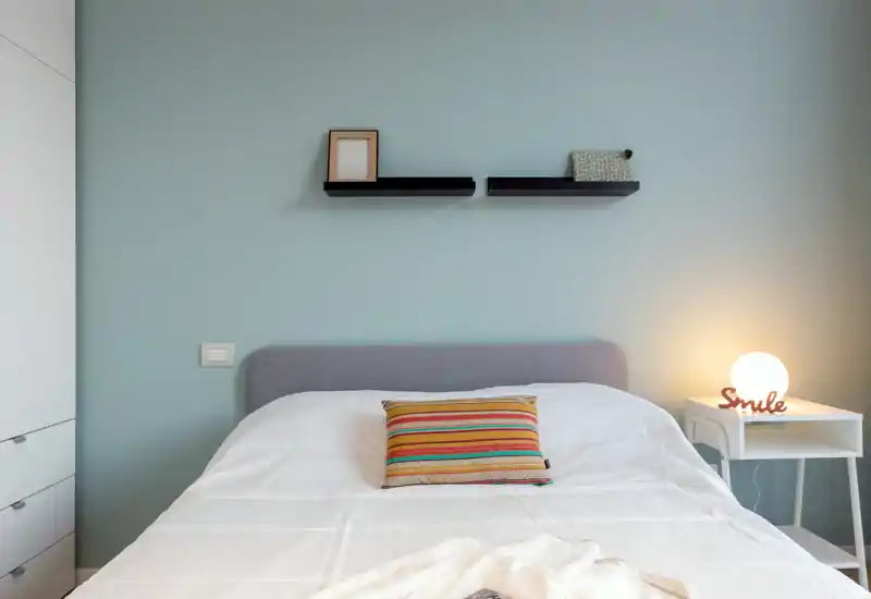 Dormitorio luminoso y minimalista con cama doble cómoda, cojín de colores y lámpara de mesita que crean un ambiente acogedor y moderno.