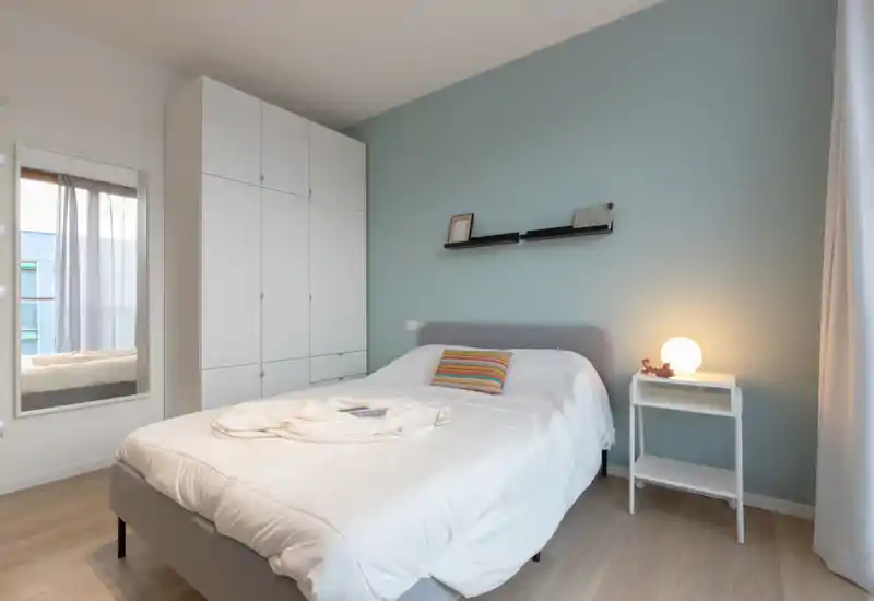 Dormitorio moderno y luminoso con cama doble bien hecha, pared azul suave, armario empotrado y mesita con lámpara cálida, ideal para una estancia cómoda.