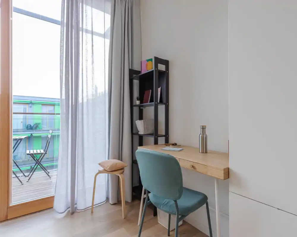 Bright living area with a compact desk and comfortable upholstered chair next to a balcony door; sheer curtains and a small shelving unit create a calm, functional workspace within the living room.