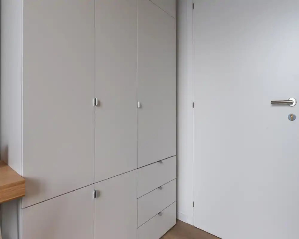 Compact, modern entry area featuring built-in white storage cabinets and a plain white interior door, with light wood flooring.