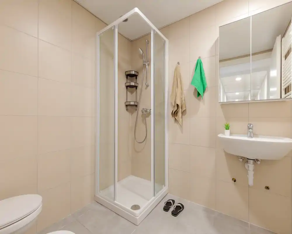 Baño moderno y luminoso con ducha de esquina de cristal, lavabo mural, espejo-armario y azulejos beige neutro — limpio y bien iluminado para huéspedes.