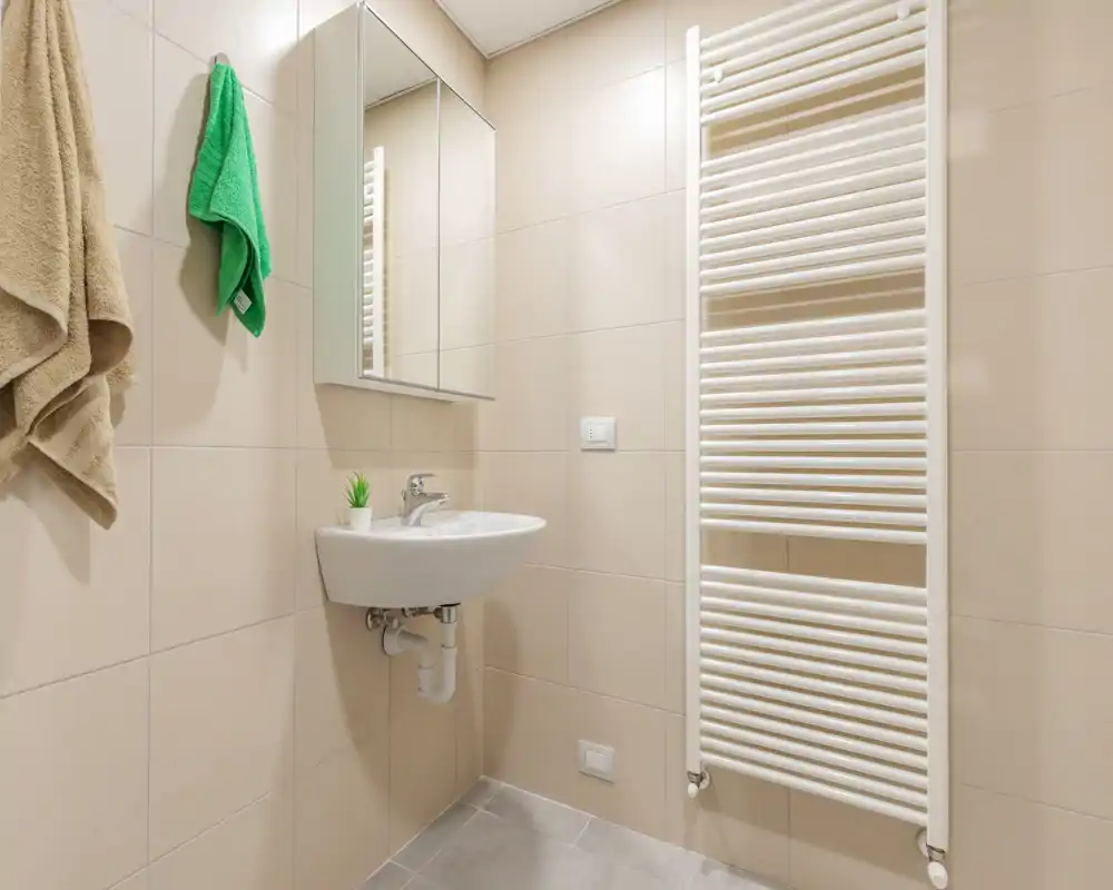 Baño moderno y compacto con lavabo suspendido, armario espejo y toallero calefactado; azulejos neutros y buena iluminación crean un espacio limpio y cuidado.