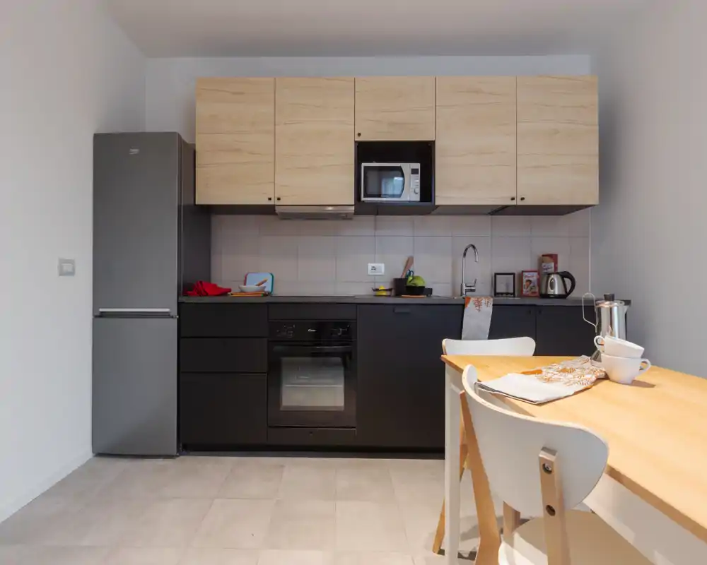 Cocina compacta y moderna con muebles bajos negros, armarios altos de madera clara, horno empotrado y frigorífico; una pequeña mesa con sillas crea un acogedor rincón para desayunar.