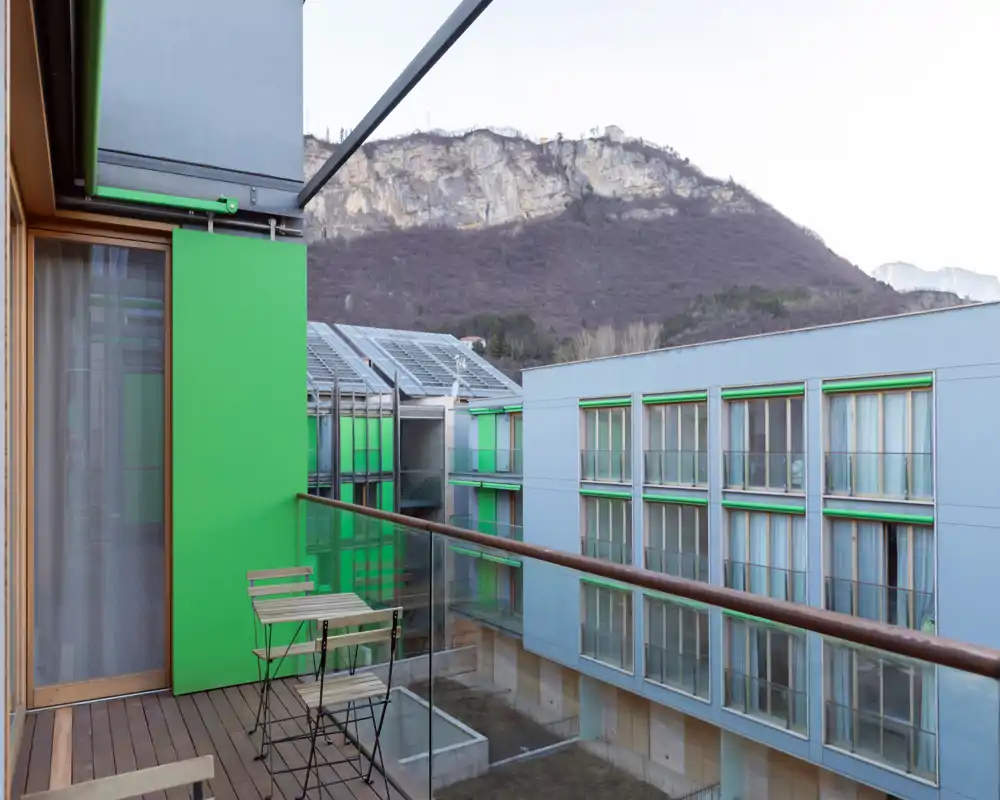Balcón soleado con dos sillas plegables y una mesa pequeña, barandilla de vidrio y vistas al patio y a la ladera rocosa cercana — ideal para el café de la mañana.