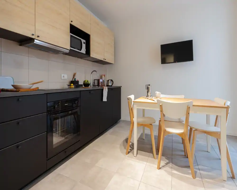Cocineta moderna y luminosa con muebles bajos negros, armarios de madera clara y una pequeña mesa para cuatro — ideal para un apartamento compacto.