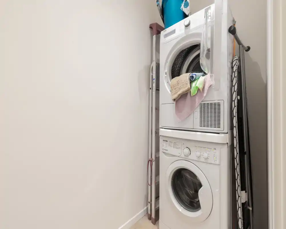Pequeño cuarto de lavandería/servicio con lavadora y secadora apiladas, tabla de planchar y escalera guardadas — espacio funcional, luminoso y bien organizado.