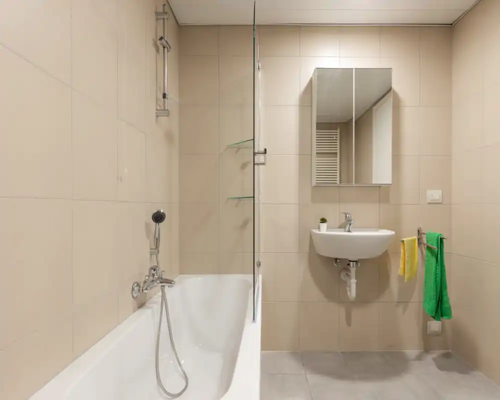 Baño moderno y limpio con bañera, lavabo mural y armario espejo; azulejos beige neutros y buena iluminación que crean un espacio luminoso y funcional.