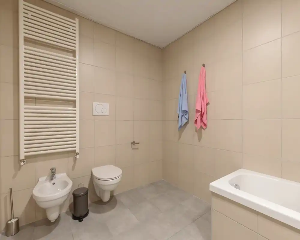 Baño moderno y espacioso con inodoro suspendido, bidé, bañera y toallero térmico; azulejos beige neutros y dos toallas de colores aportan un aspecto sencillo y fresco.