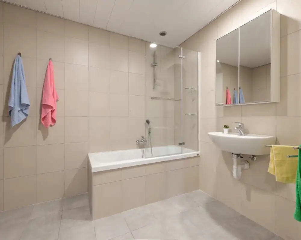Baño moderno y bien iluminado con bañera y lavabo de pared, azulejos beige y armario con espejo — limpio y funcional.