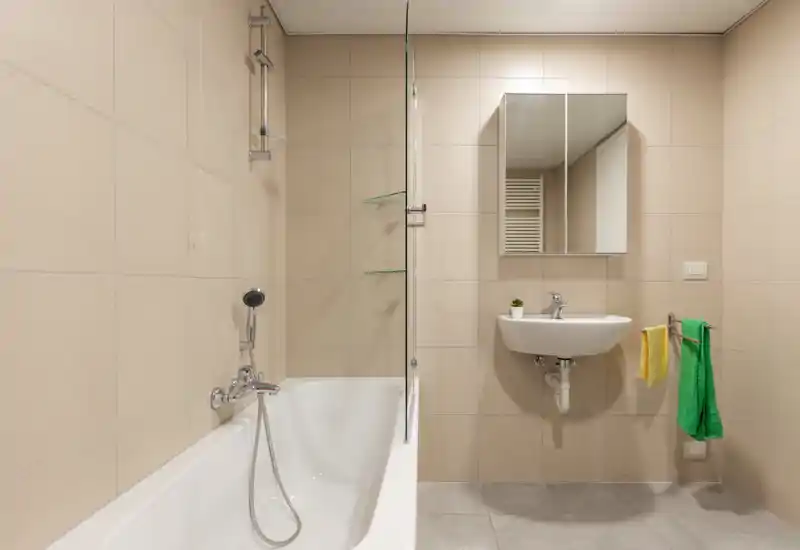 Modern, compact bathroom with a bathtub and handheld shower on the left, a wall-mounted sink and mirrored cabinet on the right, and neutral beige tiles creating a clean, minimal look.
