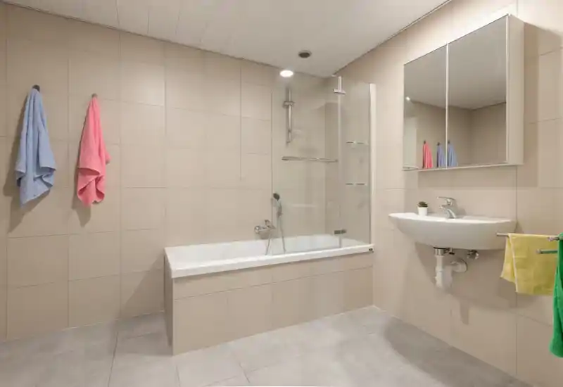 Modern, clean bathroom with a built-in bathtub, glass shower screen, wall-mounted sink and mirrored cabinet; neutral beige tiles and good overhead lighting create a bright, tidy space.