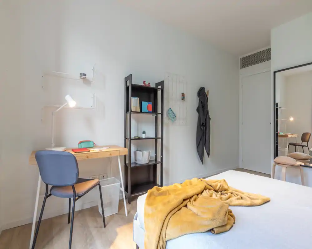Bright, modern bedroom with a neatly made bed draped in a warm mustard throw, a compact wooden desk and shelving — ideal for a comfortable stay or remote work.