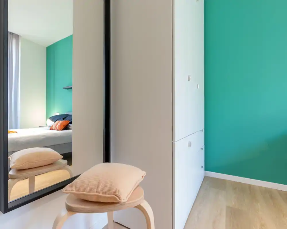 Bright, modern bedroom corner with a visible bed reflected in a large mirror, teal accent wall and simple storage units; soft cushions and natural light create a welcoming atmosphere.
