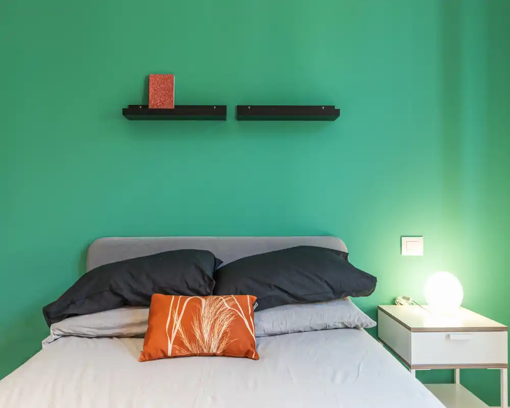 Bright, modern bedroom with a grey bed, dark pillows and an orange decorative cushion against a teal wall; bedside table with a glowing lamp creates a cozy, well-styled atmosphere.