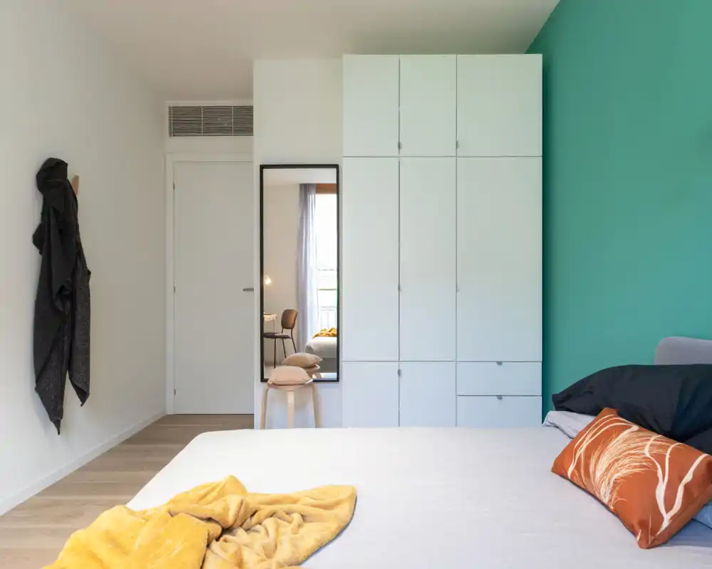 Bright, modern bedroom with a neatly made bed, teal accent wall and built-in wardrobe; soft textiles and a full-length mirror add warmth and function.