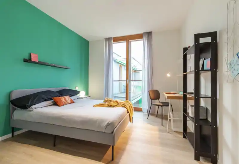 Bright, modern bedroom with a large double bed, green accent wall, natural light from a floor-to-ceiling window, a small desk and shelving — cozy and well-styled for rental listings.