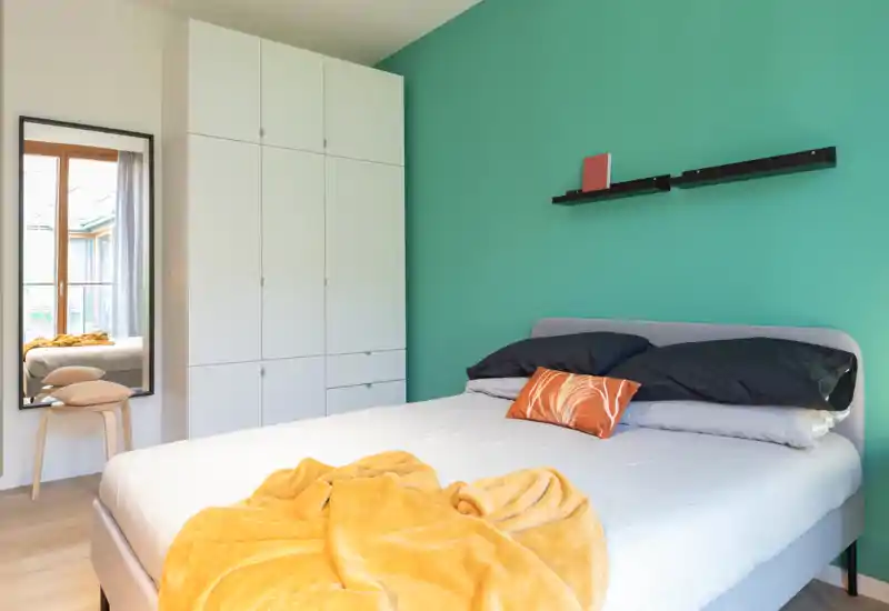 Bright, modern bedroom with a comfortable double bed against a teal accent wall, neatly arranged pillows and a cozy mustard throw. A tall white wardrobe and a full-length mirror by the window complete the minimalist, airy layout.