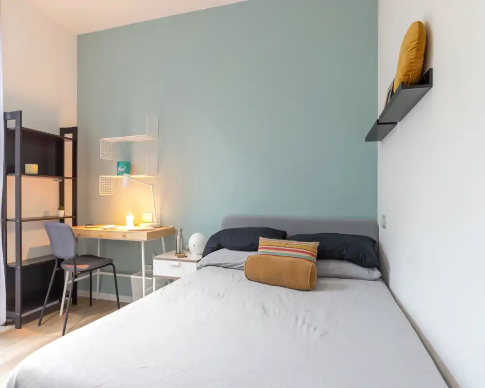 Chambre moderne et lumineuse avec un lit bien fait, coussins décoratifs et un petit bureau avec lampe d’appoint chaleureuse.