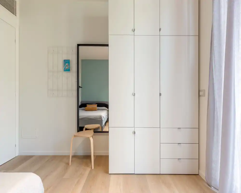 Chambre moderne et minimaliste avec une grande armoire blanche et un miroir reflétant le lit ; lumière naturelle douce et parquet chaud pour une ambiance apaisante.