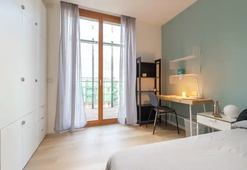 Dormitorio moderno y luminoso con la cama parcialmente visible en primer plano, un escritorio de madera con lámpara junto a una pared verde suave y una amplia puerta acristalada que da a un pequeño balcón.