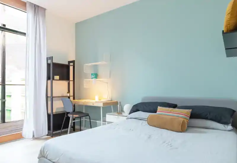 Dormitorio luminoso y moderno con una cama amplia y cómoda, ropa de cama suave y un escritorio junto a la ventana. Pared en tonos pastel, cojines decorativos y luz natural que crean un ambiente acogedor y funcional.