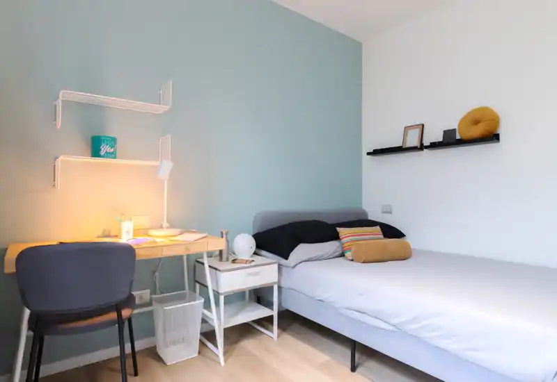 Dormitorio individual moderno y luminoso con cama bien hecha, mesita de noche y un escritorio compacto con lámpara — ideal para estudiante o ocupante individual.