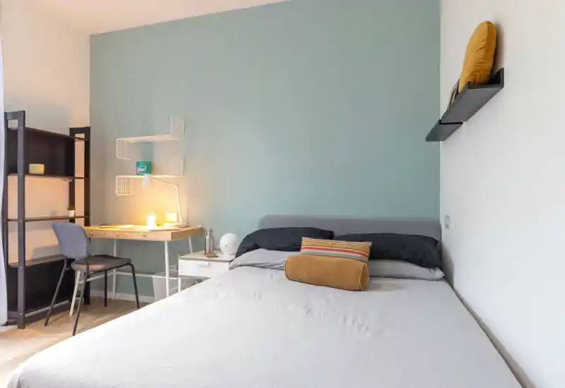 Dormitorio moderno y luminoso con cama bien hecha, cojines decorativos y un pequeño escritorio con lámpara cálida.