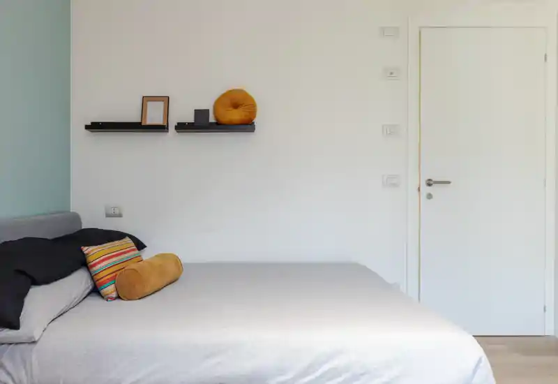Dormitorio luminoso y minimalista con una cama grande vestida en tonos neutros y cojines coloridos; estantes de pared añaden un toque moderno.