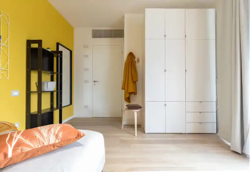 Rincón de dormitorio moderno y luminoso con la cama parcialmente visible en primer plano, pared amarilla de acento, estantería abierta y un armario blanco alto. La luz natural y los textiles suaves crean un ambiente acogedor, ideal para anuncios de alquiler.
