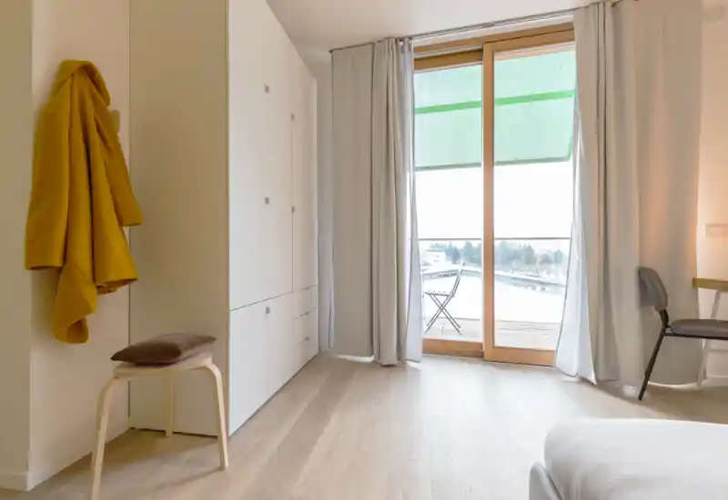 Dormitorio moderno y luminoso con luz natural desde la puerta al balcón, textiles suaves y mobiliario minimalista que crean un ambiente tranquilo y acogedor.