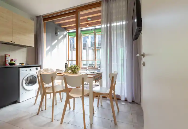 Compact modern kitchen-dining area with a small wooden table and four chairs, large glass doors leading to a balcony, and integrated appliances including a washing machine. Bright, naturally lit space with simple Scandinavian-style furniture and a potted centerpiece on the table.