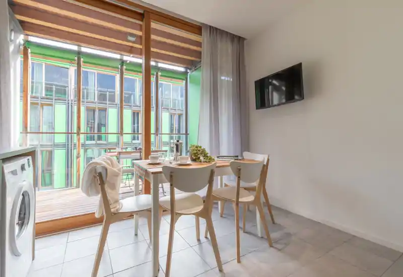 Bright, modern dining area with a small wooden table set for four, large sliding glass doors opening to a balcony and a wall-mounted TV.