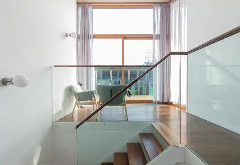 Bright, minimalist living area with two comfortable chairs placed by a large floor-to-ceiling window, hardwood floors and glass balustrade creating an airy, modern atmosphere.