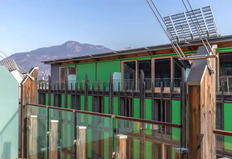 Sunny balcony/terrace view with glass balustrades, wooden posts and a green-clad neighboring building; clear mountain backdrop and pleasant daylight.