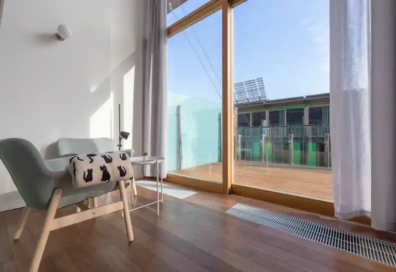 A bright, modern sitting area with a comfortable armchair, side table and large sliding glass door opening to a sunlit balcony — ideal for morning light and relaxation.