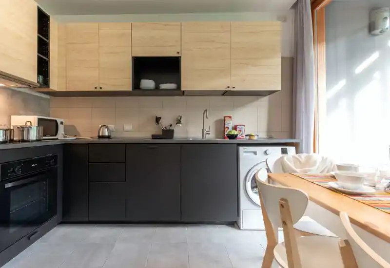 Modern kitchenette with matte dark lower cabinets, light wood upper cupboards and a small dining table by a bright window; includes oven, sink and washer — compact and functional.