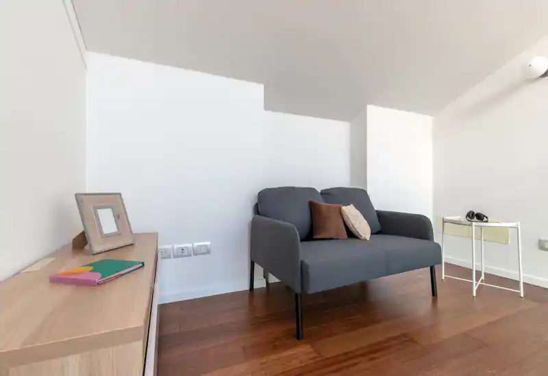 Compact, well-styled living area with a modern two-seater sofa, decorative cushions and warm wooden flooring — bright and inviting for relaxing or reading.