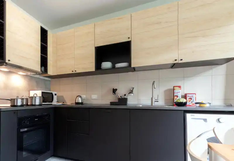 Modern kitchenette with matte black lower cabinets and light wood upper cupboards, integrated oven, countertop appliances and a washing machine visible at the right; clean, well-lit space ideal for apartment listings.